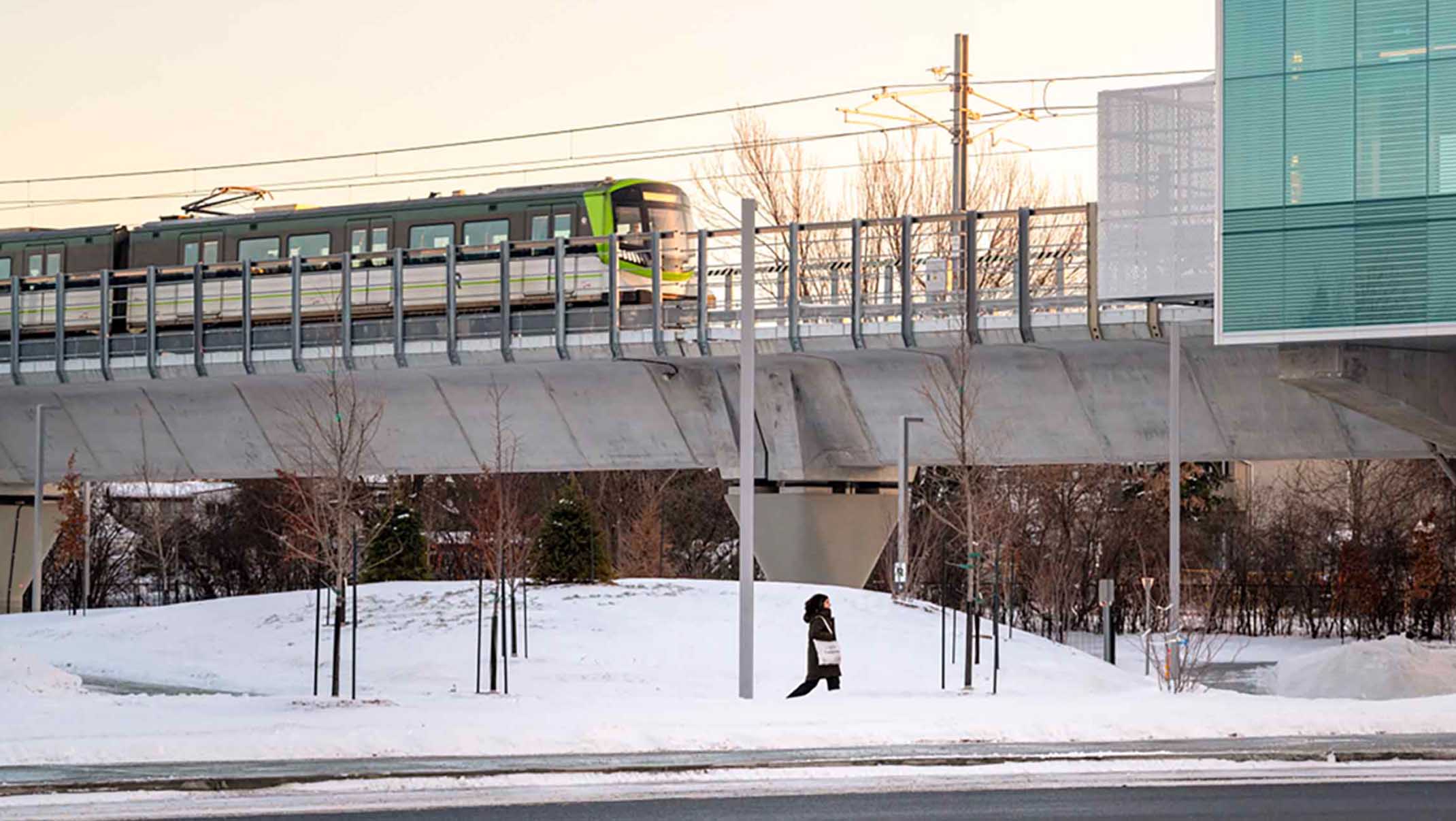 A person walking in the winter with the overhead LRT in the background