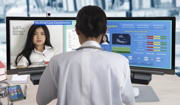 Female doctor working with two computers for distance patient consultation and searching her health information from medical record system.
