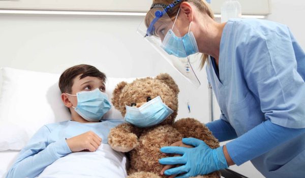 Nurse takes care of the patient child in hospital bed playing with teddy bear, wearing protective masks, corona virus covid 19 protection concept, 