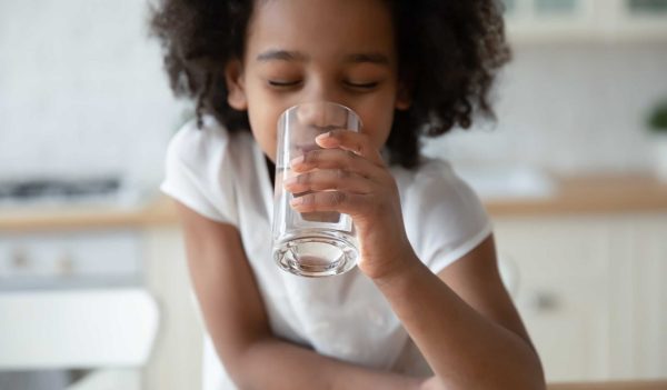 Close up head shot satisfied pretty African American little girl drinking pure water, holding glass, positive cute child kid enjoying mineral water, healthy lifestyle and refreshment concept