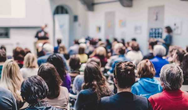 Make speaker, blurred in the background, presenting to a room full of people facig him.