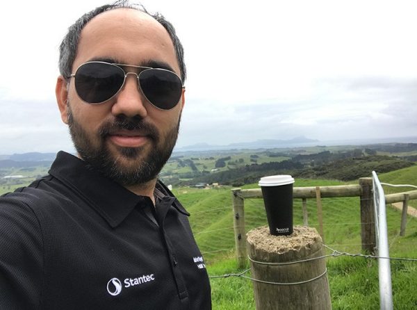 Maninder outside with a coffee cup on a post with a rural landscape in the background.